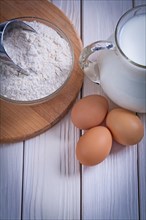 Aerial view flour in bowl scoop eggs jug with milk on white lacquered old wooden board food and drink concept