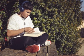 A teenager with headphones reads a book on a wooden bench