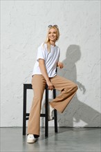 Happy adult woman in urban-style casual clothes sitting on tall stool in white studio