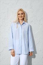 Comfortable summer outfit. Smiling woman in blue cotton blouse next to white wall