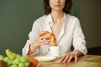Unrecognizable woman dipping croissant into cappuccino in the morning at home