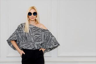 A stylish blonde woman stands indoors wearing a striped blouse and sunglasses