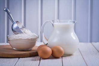 Horizontal view eggs flour in transparent bowl jug with milk on white lacquered old wooden board eating and drinking concept