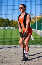 Beautiful young fitness girl warms up with a rubber expander before training at the stadium. Attractive slender brunette in an orange tracksuit. Active lifestyle