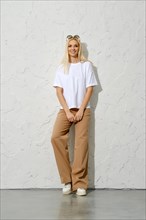 Cute smiling adult woman in oversized trousers and white t-shirt leaning on studio wall