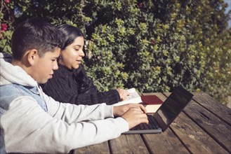 Two children studying with a laptop and books on a wooden table outdoors on a sunny day