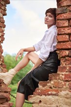 Image of a stylish beautiful woman in a white shirt and leather skirt in a park against the background of a destroyed building. The concept of style and fashion