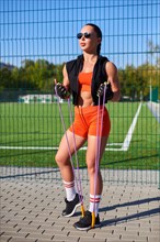 Beautiful young fitness girl warms up with a jump rope before training at the stadium. Attractive slim brunette in an orange tracksuit. Active lifestyle