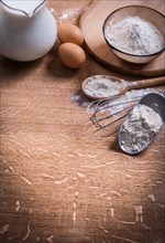 Flour in scoop Spoon and bowl Eggs Pitcher of milk on wooden board
