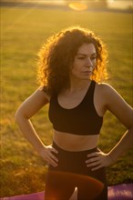 Young curly athletic girl in sportswear relaxing on a yoga mat outdoors on the grass during sunset