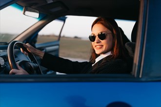 A young beautiful stylish girl driver in a jacket and sunglasses driving a car smiles and greets passengers