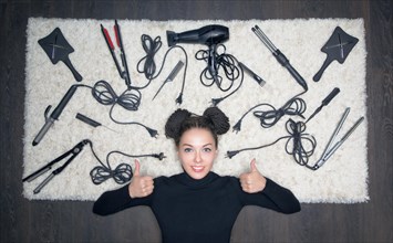 Charming girl with a smile lies on a white carpet. Her thumbs are up. Against the backdrop of the tools for creating a haircut
