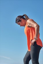 Woman in orange tank top and black leggings resting or contemplating against a clear blue sky