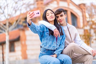 Multi-ethnic teen couple taking a selfie sitting and embracing in an urban park