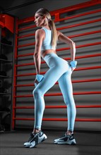 Beautiful tall blonde posing in the gym with dumbbells in her hands against the background of the wall bar. The concept of sports
