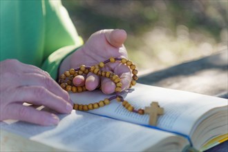 A woman's hands praying the rosary in the field over an open bible on a wooden table