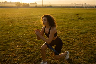 Young curly athletic girl in sportswear performs lunges with resistance band outdoors on the grass during sunset
