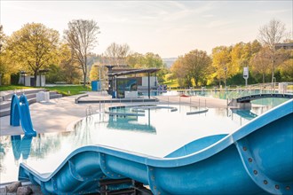 An empty outdoor pool with slide and water surface surrounded by autumnal trees under a clear sky
