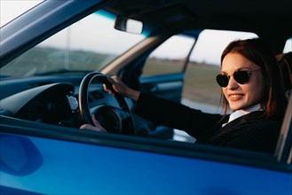 A young beautiful stylish girl driver in a jacket and sunglasses driving a car smiles and greets passengers