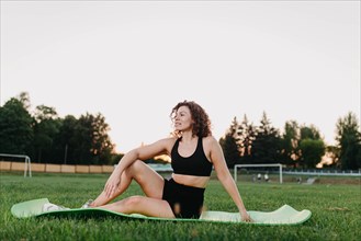 Young curly athletic girl in sportswear resting between exercises while sitting on a yoga mat outdoors on the grass during dawn