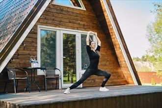 Sporty woman in sport clothing does stretching exercise on the terrace of her wooden cabin