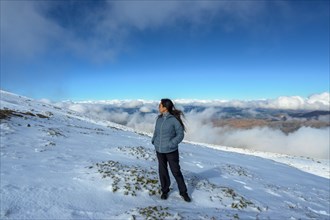 A person stands facing a snowy mountain landscape under a blue sky with clouds