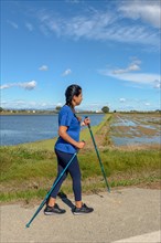 A woman in a blue shirt Nordic walking along a waterway on a sunny day with a clear blue sky