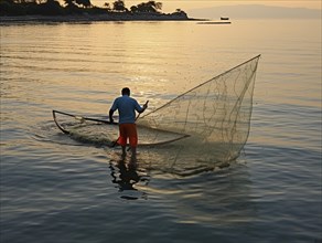 A solitary fisherman on a boat works with his net as the sunrise colors the sky peacefully