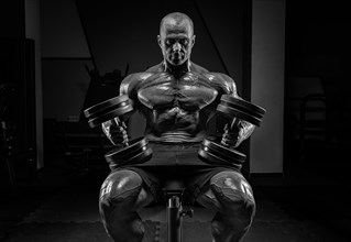 Muscular man sits on a bench with dumbbells. Bodybuilding and powerlifting concept.