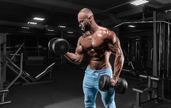 Muscular man poses in the gym in jeans with dumbbells in his hands. Sports