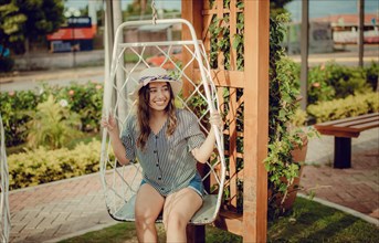 Relaxed girl in hat sitting on a white swing in a beautiful garden. A smiling girl sitting on a swing in La Calzada
