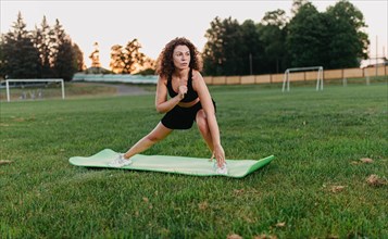 Young curly athletic girl in sportswear performs leg stretching on a yoga mat outdoors on the grass during sunrise