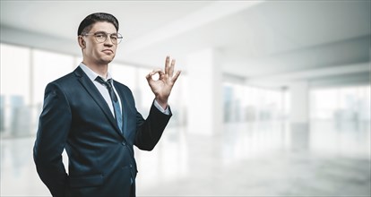 Portrait of a businessman in a suit. He is standing in the office of a skyscraper. Ok sign. Business concept.