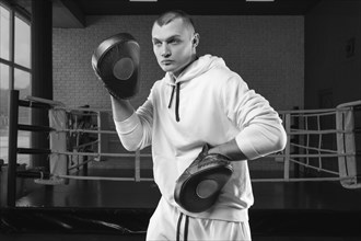 Male trainer in the gym against the background of the ring holds boxing paws. Mixed martial arts concept. High image quality