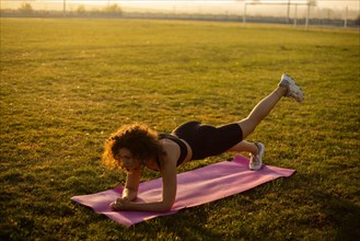 Young curly athletic girl in sportswear performs a buttock exercise on a yoga mat outdoors on the grass during sunset