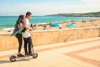 A couple on a scooter enjoying a sunny day at the beach with people and the sea in the distance
