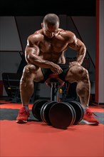 Muscular man sits on a bench near huge dumbbells. Bodybuilding and powerlifting concept.