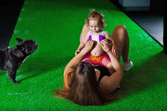 Charming sports mom plays in the gym with her little daughter and a French bulldog.