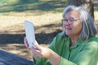 Mature white-haired woman with glasses on a video call with her tablet in the countryside on a sunny day