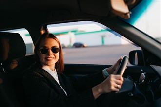 A young beautiful stylish girl driver in a jacket and sunglasses behind the wheel of a car greets passengers