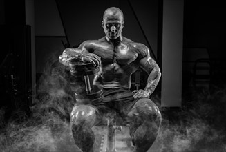 Muscular man sits on a bench in smoke with dumbbells. Bodybuilding and powerlifting concept.