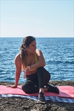 A woman sits on a yoga mat by the ocean