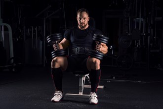 Impressive athlete sits on a bench in the gym with two dumbbells on his legs