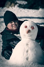 Boy lying in the snow next to self-made snowman with carrot nose and tomato eyes