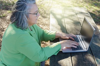 Mature woman with white hair and glasses working on her computer in the sunlit countryside
