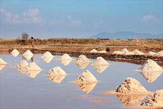 Salt piles symmetrically reflected in the water under a clear blue sky with a person in the distance. Kampot