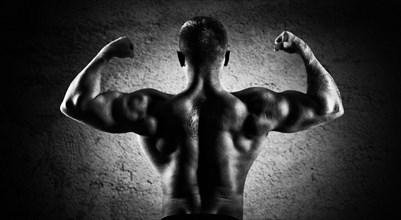 Huge weightlifter is standing with his back to the camera and straining his hands demonstrating a double bicep. Back view