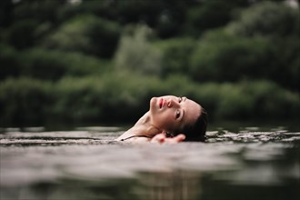 A young slender beautiful and hot girl with wet hair and big lips swims in the lake