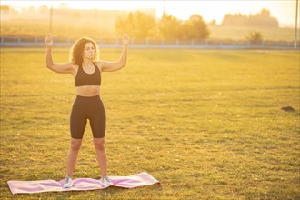 A young curly athletic girl in sportswear doing a warm-up on the upper shoulder girdle in the fresh air during sunset