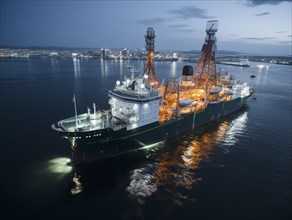 Illuminated offshore drilling ship at night with a cityscape in the background